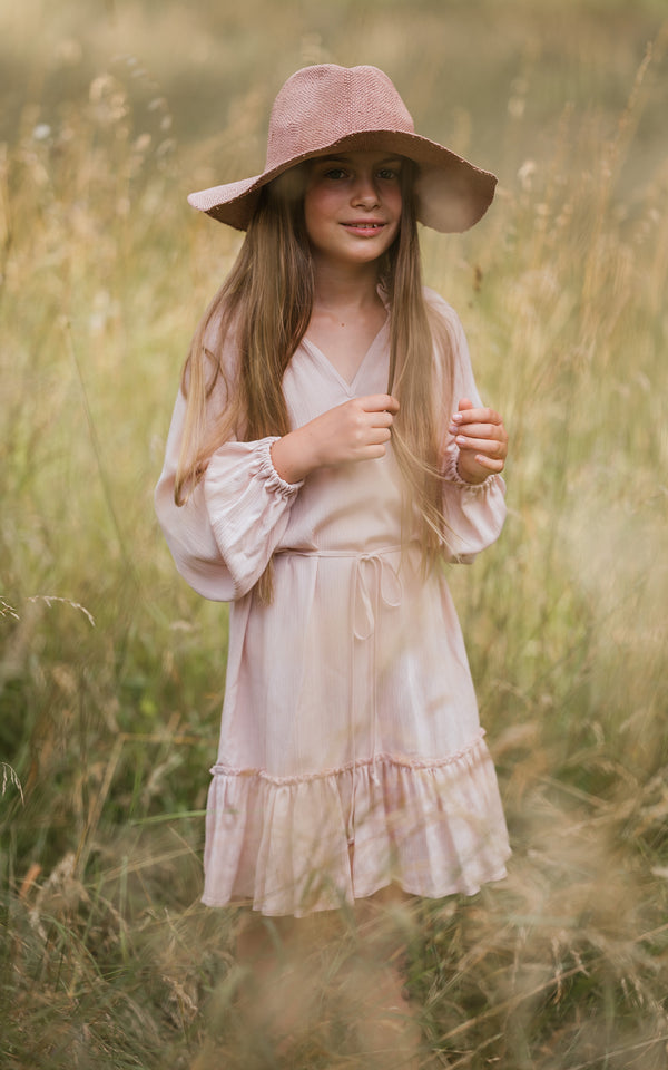 Das Mädchen trägt ein luftig leichtes knielanges Sommerkleid aus Viskose in zartem Rosé und trägt dazu einen Boho-Style Strohhut. Carmensita | Sommerkleider made in Austria.